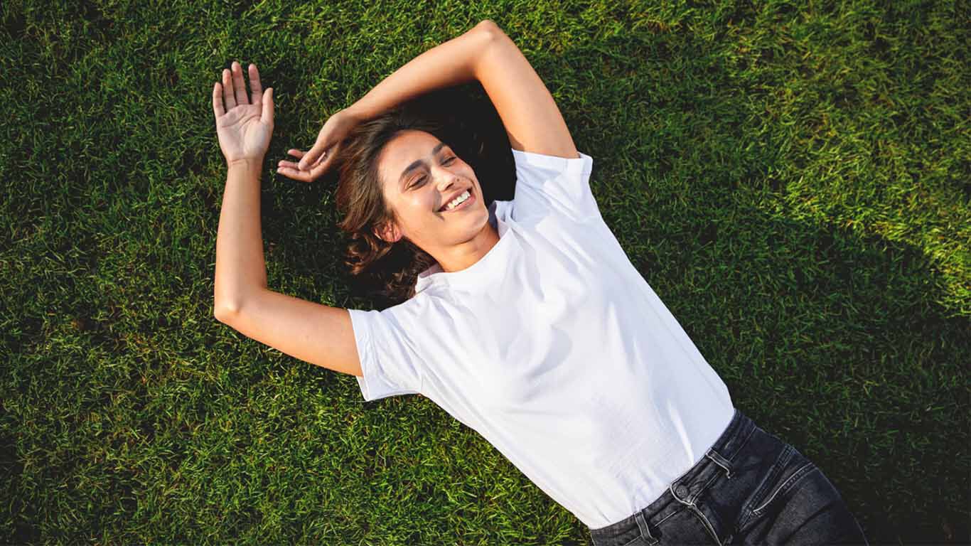 Une femme aux cheveux fonc&eacute;s est allong&eacute;e, d&eacute;tendue et souriante, dans un pr&eacute; verdoyant